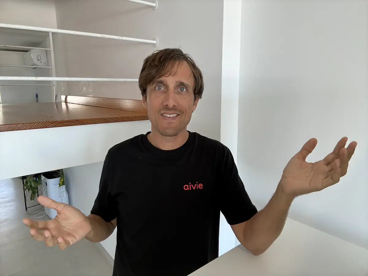 Adrian Schimpf im schwarzen Aivie-T-Shirt hebt fragend beide Hände